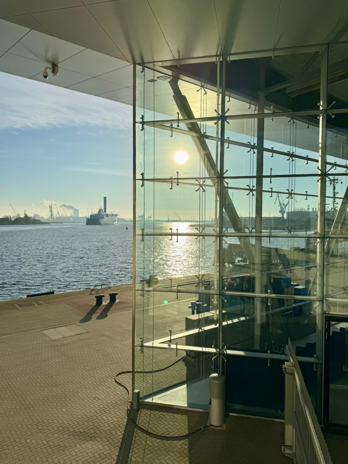 Detailaufnahme des Kreuzfahrthafens Warnemünde mit Blick Richtung Seehafen: Eine Fähre fährt auf dem Seekanal, während die Sonne durch die Glasfenster des Terminalgebäudes P8 im Vordergrund scheint.