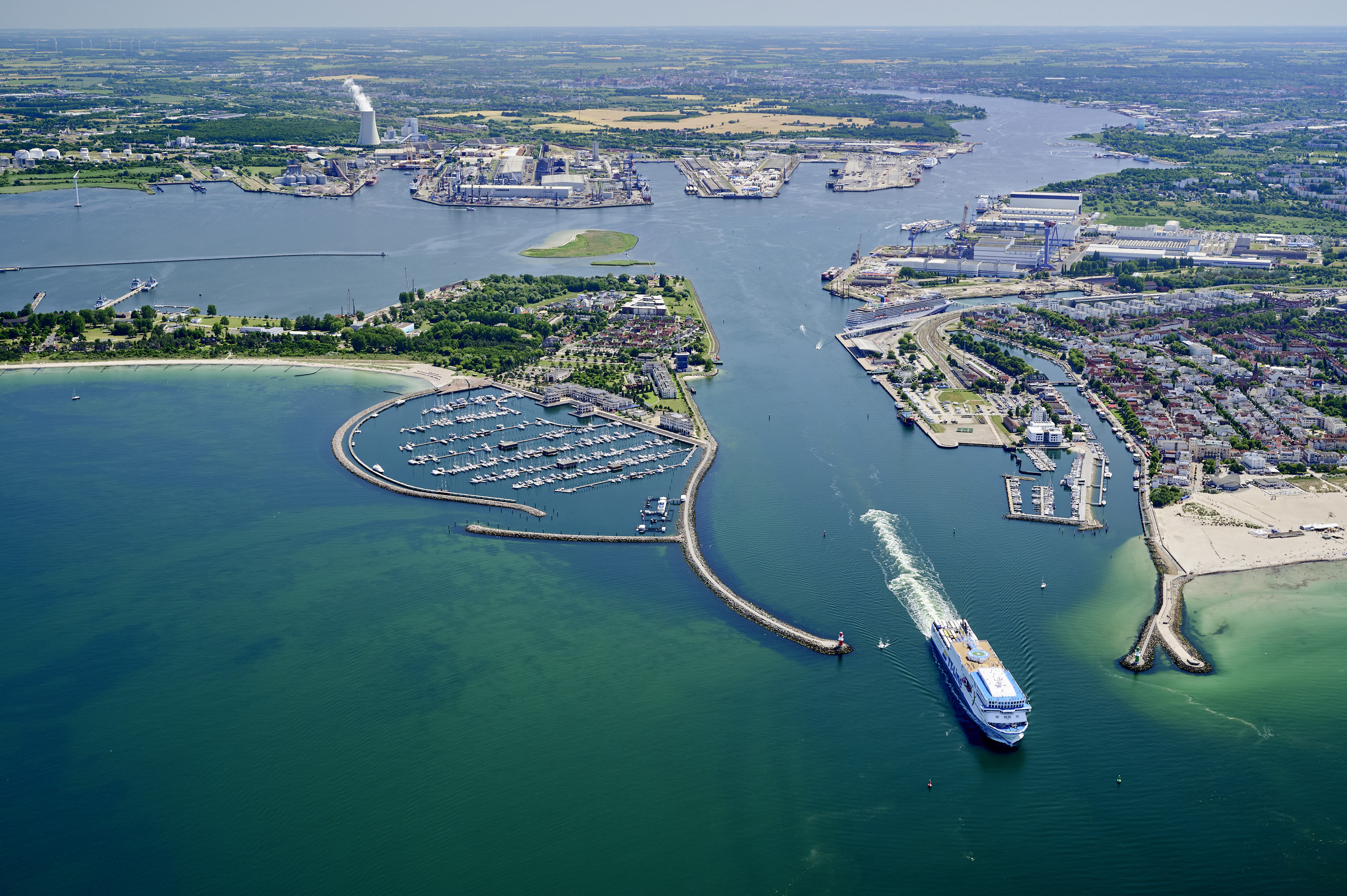 Ein Fährschiff fährt durch die Warnemünder Molen; im Hintergrund liegt ein Kreuzfahrtschiff am Terminal und der Rostocker Seehafen ist zu sehen.