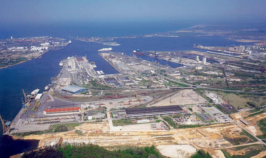 Luftaufnahme des Überseehafens Rostock mit Kaianlagen, Logistikflächen und Schiffen vor der Ostsee. Im Vordergrund liegen Felder und Wohngebiete.