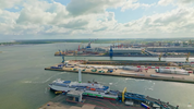Luftbildaufnahme des Überseehafen Rostock aus westlicher Richtung. Vorne am Fährterminal liegt eine Fähre mit Flettner-Rotor, auf die Autos hinaufahren. Auf dem Massengutterminal liegen Teile von erneuerbaren Energiegewinnung. Die Produktionsfläche von Liebherr und das Getreideterminal erstrecken sich bis zum Horizont. Am Himmel sind Wolken.