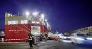Ein großes rotes Autotransportschiff namens „Malacca Highway“ liegt nachts im beleuchteten Rostocker Seehafen, während Neufahrzeuge über eine Rampe von Bord fahren.
