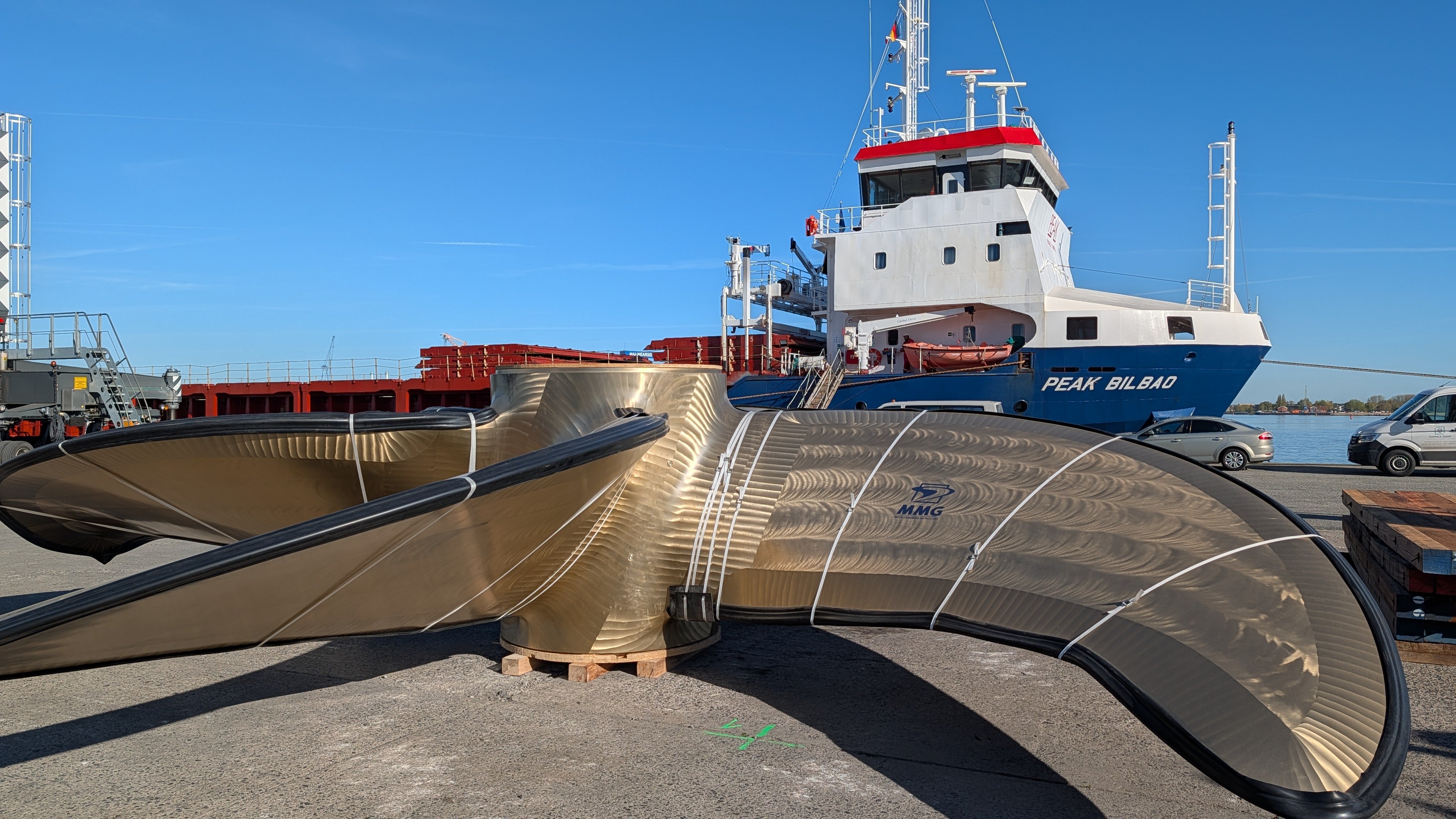 Eine große, goldglänzende Schiffsschraube lagert auf der Pier im Stückgutterminal des Rostocker Seehafens; im Hintergrund liegt ein Frachtschiff vor blauem Himmel.