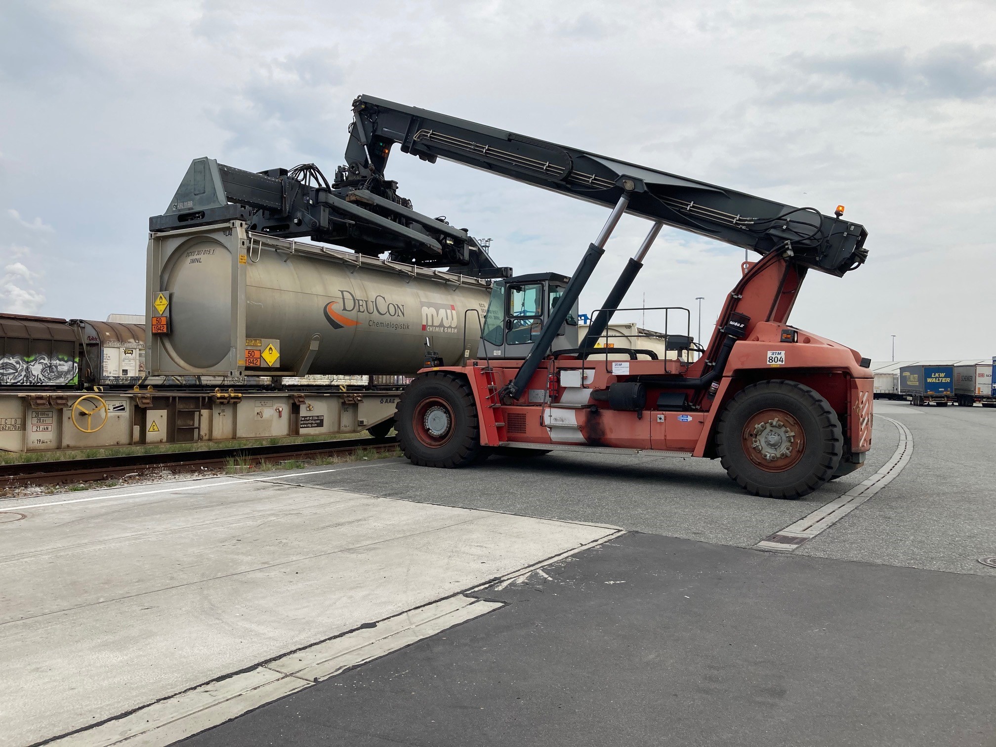 Ein Reachstacker hebt auf dem Rostocker Terminal für kombinierten Ladungsverkehr einen Tankcontainer von einem Güterzug, während im Hintergrund abgestellte Sattleauflieger zu sehen sind.