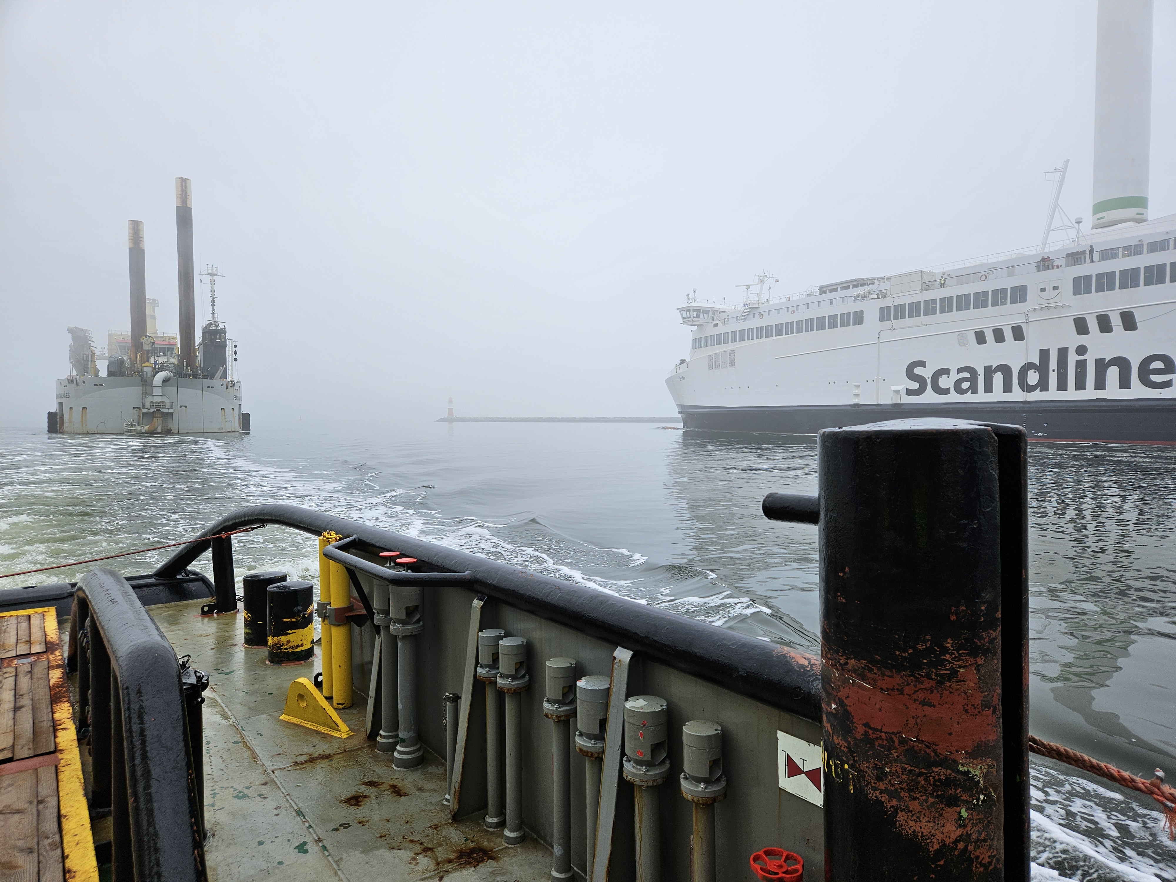 Ein großes, spezialisiertes Baggerschiff (Schneidkopfmutterbagger) namens „Fernão de Magalhães“ der Firma Jan De Nul (DJN) liegt bei nebligem Wetter im Seekanal von Rostock. Auf der rechten Bildseite fährt ein Fährschiff der Scandlines an dem Arbeits- und Baggerschiff vorbei.