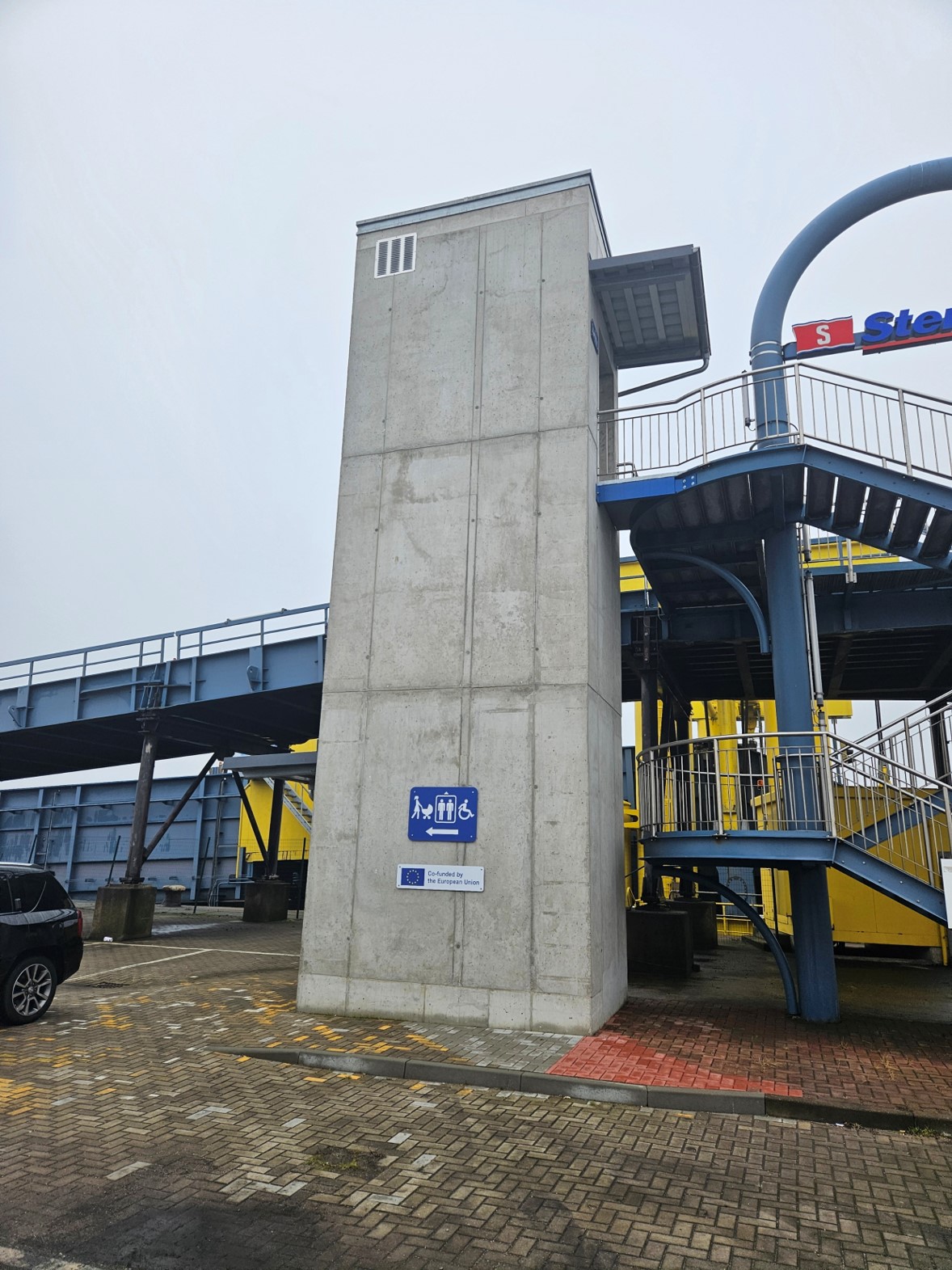 Außenansicht des neuen Beton-Fahrstuhls am Liegeplatz 64 auf dem Fährterminal Rostock: Die Fußgänger-Gangwaybrücke ist oben am Fahrstuhlturm befestigt, im Hintergrund verläuft die Rampe für Autos und Lkw.