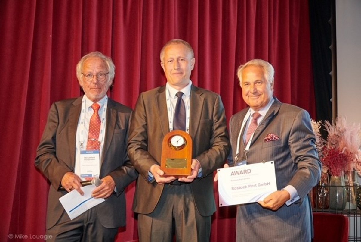 Drei Männer in Businesskleidung stehen lächelnd auf einer Bühne und blicken in die Kamera. Der Mann in der Mitte hält eine Plakette, während der rechte Mann eine Auszeichnung mit der Aufschrift „Award ROSTOCK PORT GmbH“ präsentiert.