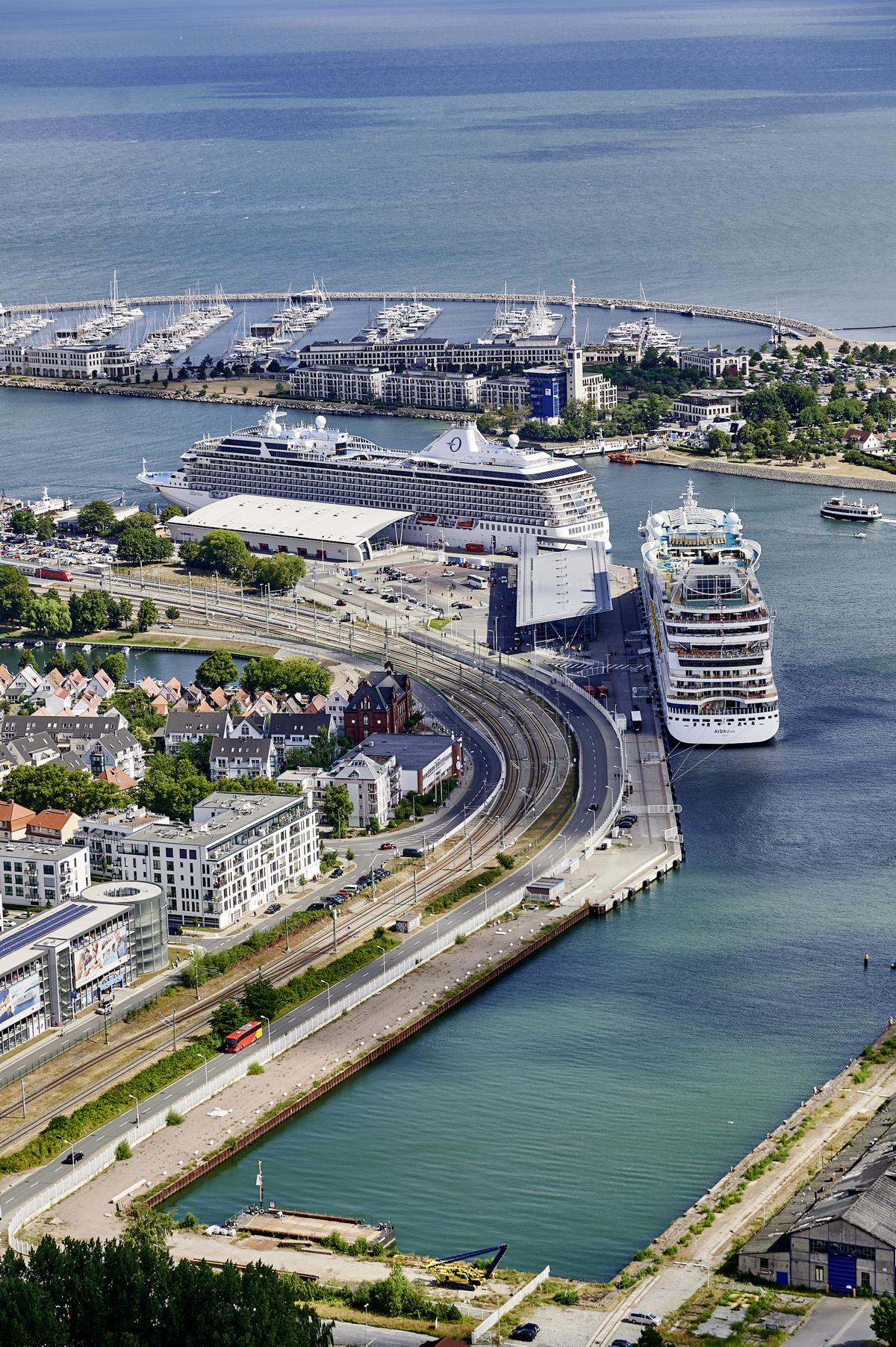 Luftaufnahme vom Kreuzfahrtterminal in Warnemünde aus Richtung des Hafenbeckens. Zwei Kreuzfahrtschiffe liegen an. Auf dem Wasser wendet ein Fahrgastschiff. Dim Yachthafen Hohe Düne liegen viele Segelboote.