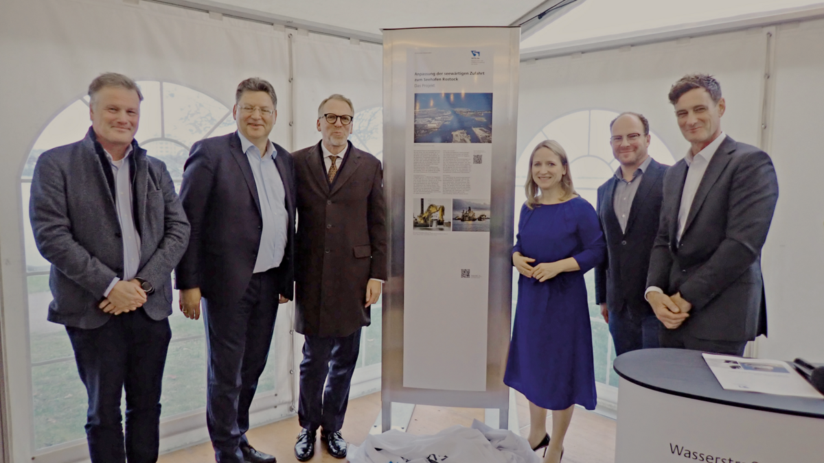 Gruppenaufnahme von sechs Personen in Business-Kleidung, die lächelnd in einem Veranstaltungszelt stehen und direkt in die Kamera blicken. In der Mitte der Gruppe ist ein Roll-up-Display aufgestellt, das Informationen zum Großprojekt der Seekanalvertiefung Rostock zeigt.